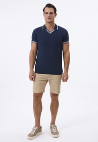 Marineblaues Poloshirt mit weißen Kragen und gestreiften Ärmeln, kombiniert mit beigen Shorts. Modell trägt graue Sneaker und steht vor einem schlichten Hintergrund.