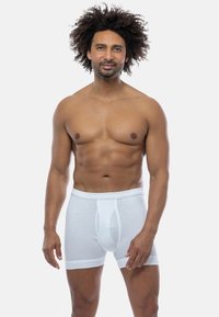 Uomo senza maglietta con capelli ricci, che indossa boxer bianchi, in piedi davanti a uno sfondo grigio chiaro uniforme.