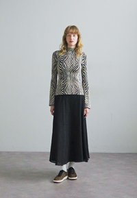 Lang svart plissert skjørt i kombinasjon med en tettsittende topp i svart-hvitt mønster med bølgete linjer. Modell iført svarte joggesko med leopardmønster.