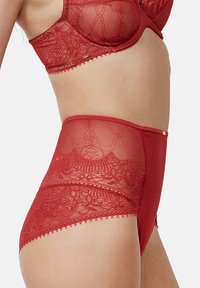 Culotte rouge taille haute avec détails en dentelle, devant lisse, bords festonnés et petits accents métalliques sur le côté.