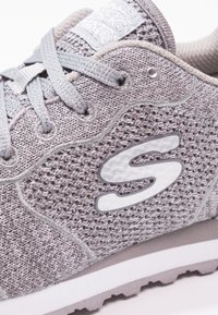 Grå atletisk sko med en texturerad mesh-överdel, silverdetaljer, traditionellt snörningssystem och en vit gummisula. Anmärkningsvärd "S"-logotypdetalj.