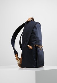 Marinblå nylonryggsäck med beige läderdetaljer, med en framficka med dragkedja och justerbara remmar, som visar en slank och strömlinjeformad design.