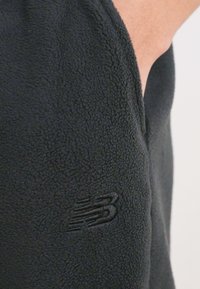 Melnā flīsa jaka ar teksturētu virsmu un diskrētu iespiestu New Balance logotipu uz piedurknes. Vienkāršs, funkcionāls dizains.