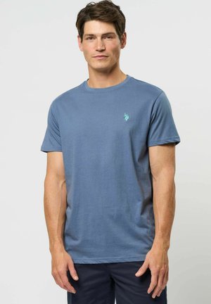 Mężczyzna ubrany w niebieski t-shirt z krótkim rękawem i okrągłym dekoltem z małym turkusowym logo na lewej piersi, stojący na neutralnym tle.