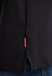 Zwart shirt met korte mouwen van structuurstof, met zijsplitten en een rood label met "JOOP! JEANS" op de onderste linkerkant.