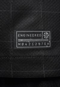 Etichetta in tessuto nera con testo bianco. Riporta "ENGINEERED" e "M0421297EH," con un simbolo di fiore. Sfondo testurizzato con motivo a griglia.