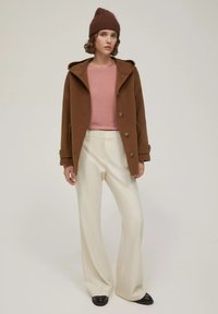 Cappotto di lana marrone con cappuccio, maglione rosa e pantaloni bianchi a gamba larga. Il modello indossa un berretto lavorato a maglia marrone e scarpe nere. Dettagli di design semplici visibili.