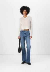 White, cropped knit sweater with a V-neck, paired with light blue flared jeans. Model holds a black bag, standing against a white background.