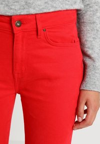 Jeans de mezclilla rojos con cintura alta, cinco bolsillos y detalle de botón de metal. Textura suave y diseño ajustado.