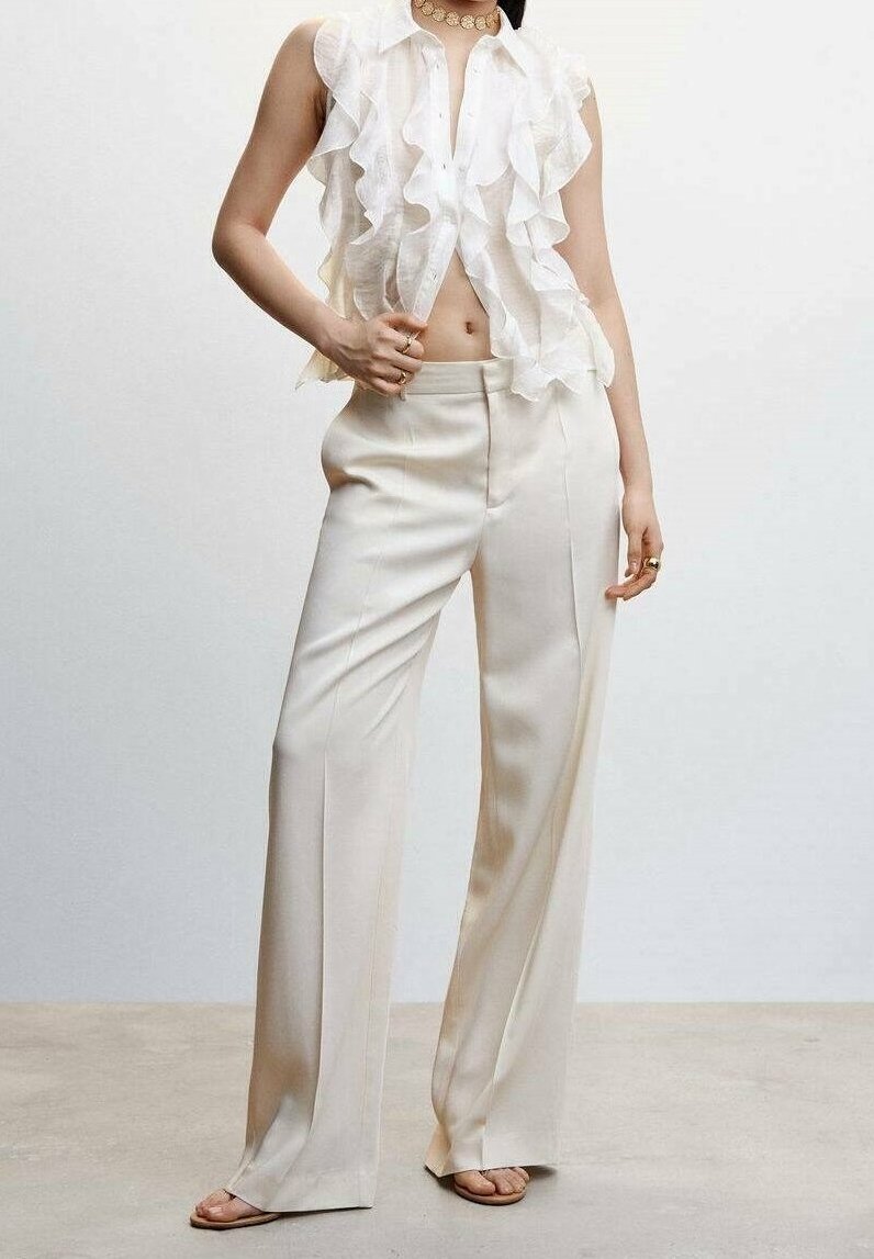 Femme portant un chemisier blanc sans manches à volants et un pantalon large crème taille haute, debout pieds nus sur un fond uni.