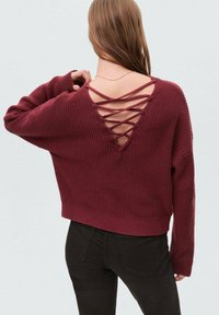 Pulover tricotat de culoare burgundă, cu o croială lejeră, având un decolteu în V adânc la spate decorat cu șnur și mansete și tiv cu aspect canalat. Purtați-l cu pantaloni negri.