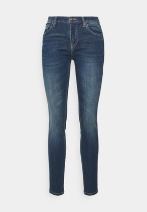 Tummansiniset slim-fit farkut, joissa on kevyt kulutus, etupainike, vetoketjukiinnitys ja viisi taskua valkoista taustaa vasten.