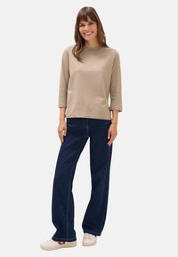 Frau steht lächelnd da und trägt ein beige-weiß gestreiftes 3/4-Ärmel-Oberteil, dunkelblaue weit geschnittene Jeans und weiße Sneakers mit roten Akzenten.