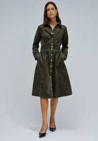 Robe chemise vert olive en doux suède synthétique, avec une taille ceinturée, un devant à boutons, des manches longues et des poches latérales. Escarpins noirs à talons pointus.