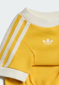 Chemise jaune à manches courtes avec col en côte blanche, poignets blancs, trois bandes blanches sur l’épaule et logo trèfle blanc sur la poitrine.