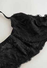 Zwarte kant bralette met transparant bloemenpatroon, geschulpte randen en verstelbare bandjes. Textuurstof voegt diepte en interesse toe.