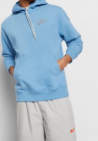 Ljusblå hoodie i mjukt material, med en framficka, dragskohet och en liten reflektiv logotyp på bröstet.