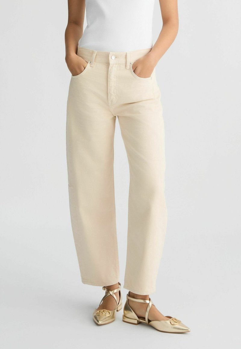 Femme portant un pantalon beige à jambes larges, les mains dans les poches, et des ballerines dorées à bout pointu avec bride autour de la cheville, sur fond uni.