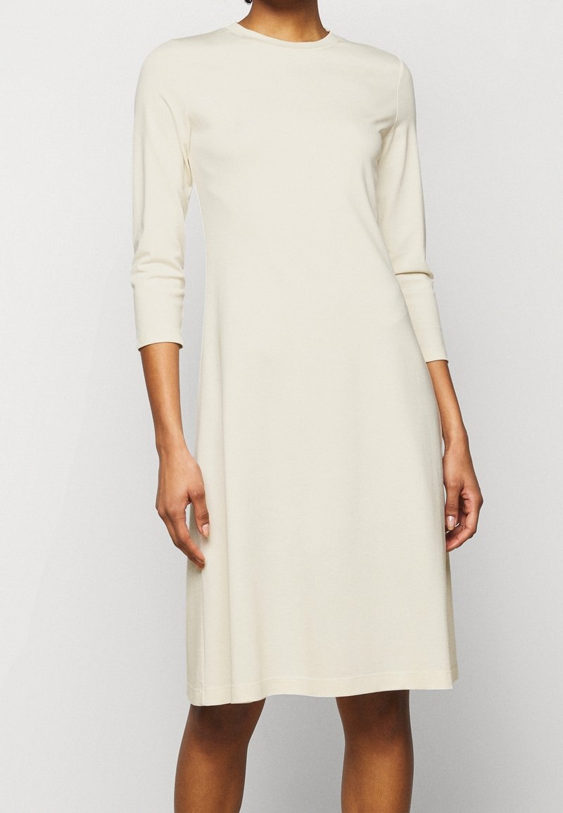 Femme portant une robe crème mi-longue, à manches longues, avec un col rond, debout devant un fond blanc uni.