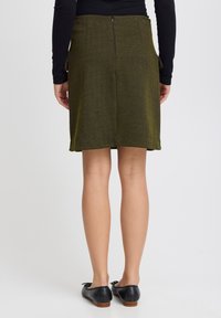 Jupe vert olive avec un motif en chevrons, longueur genou, dotée d'une fermeture éclair à l'arrière et de poches latérales. Associée à des ballerines noires.