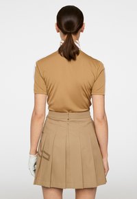 Brązowa koszulka polo z białymi akcentowanymi rękawami, połączona z jasnobeżową spódnicą z zakładkami z bocznym nadrukiem logo i zapięciem na suwak z tyłu.