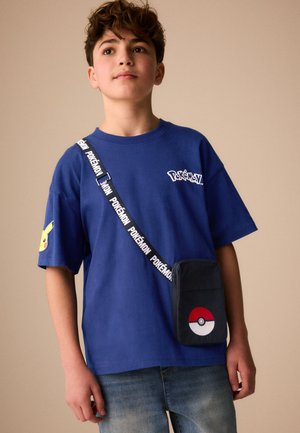 Ung person iført en blå Pokémon-t-shirt med Pikachu på ærmet og en sort crossbody taske med et Poké Ball-design.