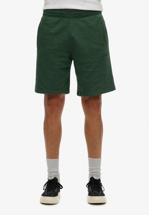 Mężczyzna w ciemnozielonych, sięgających do kolan szortach, szarych skarpetkach i czarnych trampkach, stojący na tle jednolitego białego tła.