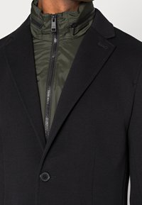 Svart kavaj med hög krage och en olivgrön zip-up jacka under. Har en slät tygkvalitet, en enda svart knapp och subtila sömnadsdetaljer.