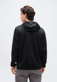 Νέος άνδρας που φοράει μαύρο μπουφάν με κουκούλα και γκρι παντελόνι, όρθιος και γυρισμένος προς τα πίσω σε απλό ανοιχτόχρωμο φόντο.