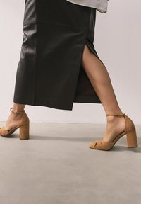 Hellbraune Wildleder-Schuhe mit hohen Absätzen und einem dicken Blockabsatz, ausgestattet mit überkreuzten Details und einem Knöchelriemen, kombiniert mit einem schwarzen Lederrock.