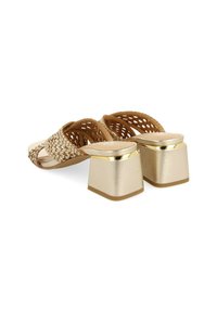Zapato tejido beige con un diseño de tira cruzada, que presenta un tacón grueso con detalles dorados y una textura suave, con una suela robusta para mayor apoyo.