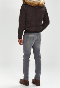 Mørkebrun bomberjakke med pelsforet hætte, båret over grå slim-fit jeans og brune støvler. Jakken har polstrede skuldre og ribbede manchetter.