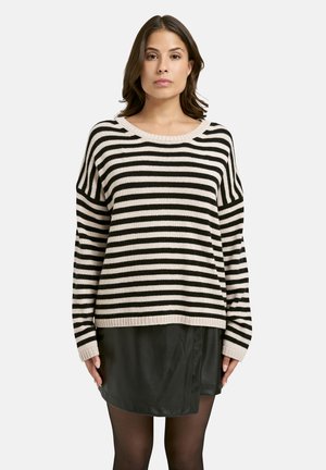 Frau, die mit Blick nach vorne in einem schwarz-weißen gestreiften Pullover und einem schwarzen Rock mit schwarzen Strumpfhosen vor einem schlichten Hintergrund steht.