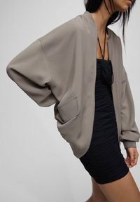 Cazadora bomber de gran tamaño en color beige con una textura suave, puños acanalados y un bolsillo con parche, superpuesta sobre un vestido negro ajustado con detalles fruncidos.