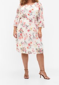 Robe fleurie avec manches courtes évasées et plissées. Base crème clair avec motifs floraux roses et verts. Portée avec des sandales à talons hauts marron à brides.