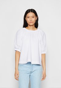 Blusa de algodão branca com um decote franzido e mangas curtas bufantes. Corte descontraído, combinada com jeans azul claro. Textura suave.