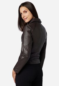 Braune Leder-Motorradjacke mit gesteppten Schulterakzenten, figurbetontem Schnitt und leicht verkürzter Länge. Glatte Textur und klassisches Kragen-Design.