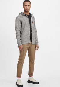Gray textured zip-up hoodie with hood, black inner lining, and brown accents. Paired with rolled brown pants and beige sneakers.