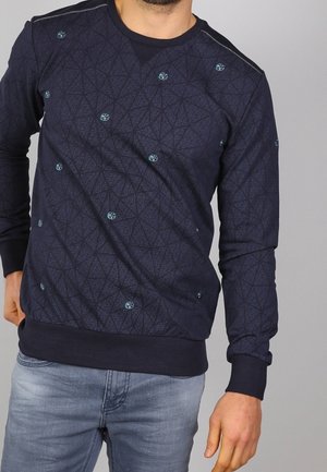 Mężczyzna ubrany w ciemnoniebieski sweter z drobnym wzorem jasnoniebieskich kółek, w połączeniu z jasnoniebieskimi dżinsami, stojący na tle szarego tła.