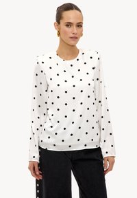 Blouse à manches longues en satin blanc à pois noirs. Présente un col rond et une coupe décontractée. Portée avec un pantalon noir.