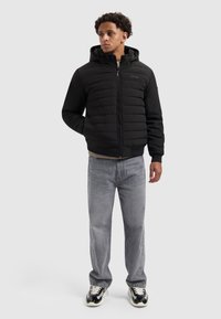 Zwarte gewatteerde pufferjack met een doorgestikte ontwerp, voorkant met rits en ribbels aan de mouwen, gecombineerd met lichtgrijze spijkerbroek en zwarte sneakers.