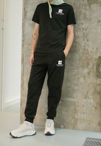 Svart bomullströja med en logotyp på vänster bröst, ihop med matchande svarta tapered joggers och ljusa sneakers. Grön scarfdetalj.