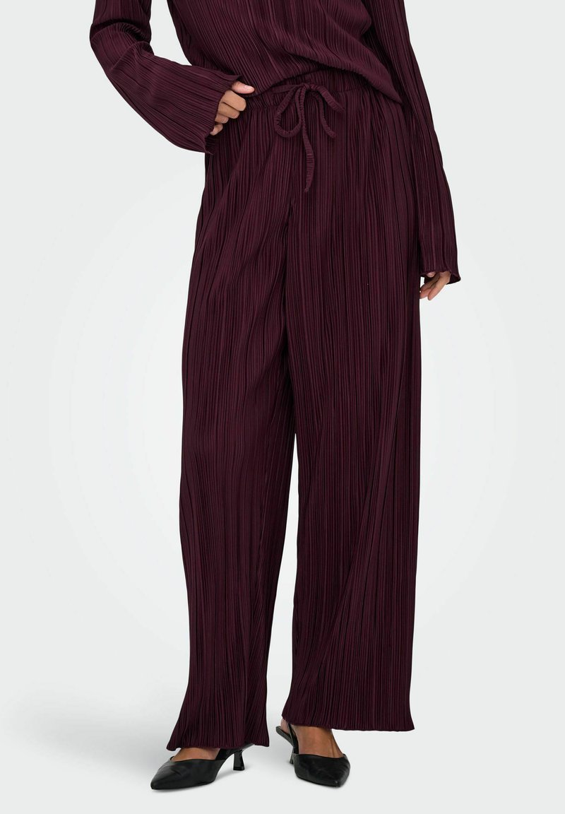 Pantalons larges plissés bordeaux avec une taille élastique et un détail de cordon de serrage, fabriqués à partir d'un tissu texturé, portés avec des talons noirs.