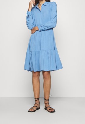 Hellblaues Hemdblusenkleid mit langen Ärmeln, geknöpftem Kragen, strukturiertem Stoff und einer gestuften Rüschensaum. Kombiniert mit schwarzen Riemchensandalen.