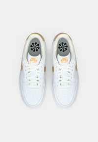 Bijele Nike Air Force 1 tenisice s glatkom kožnom gornjom delenjem, zlatnim akcentima, crnim cvjetnim ulošcima i bijelim vezicama. Okrugli vrh, perforirani dizajn.