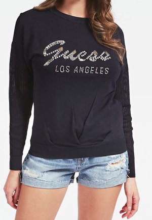 Kvinde iført sort langærmet top med "Guess Los Angeles" i rhinsten og lyseblå slidte denimshorts.