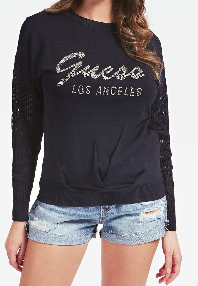 Femme portant un haut noir à manches longues avec l'inscription "Guess Los Angeles" en strass et un short en jean bleu clair déchiré.