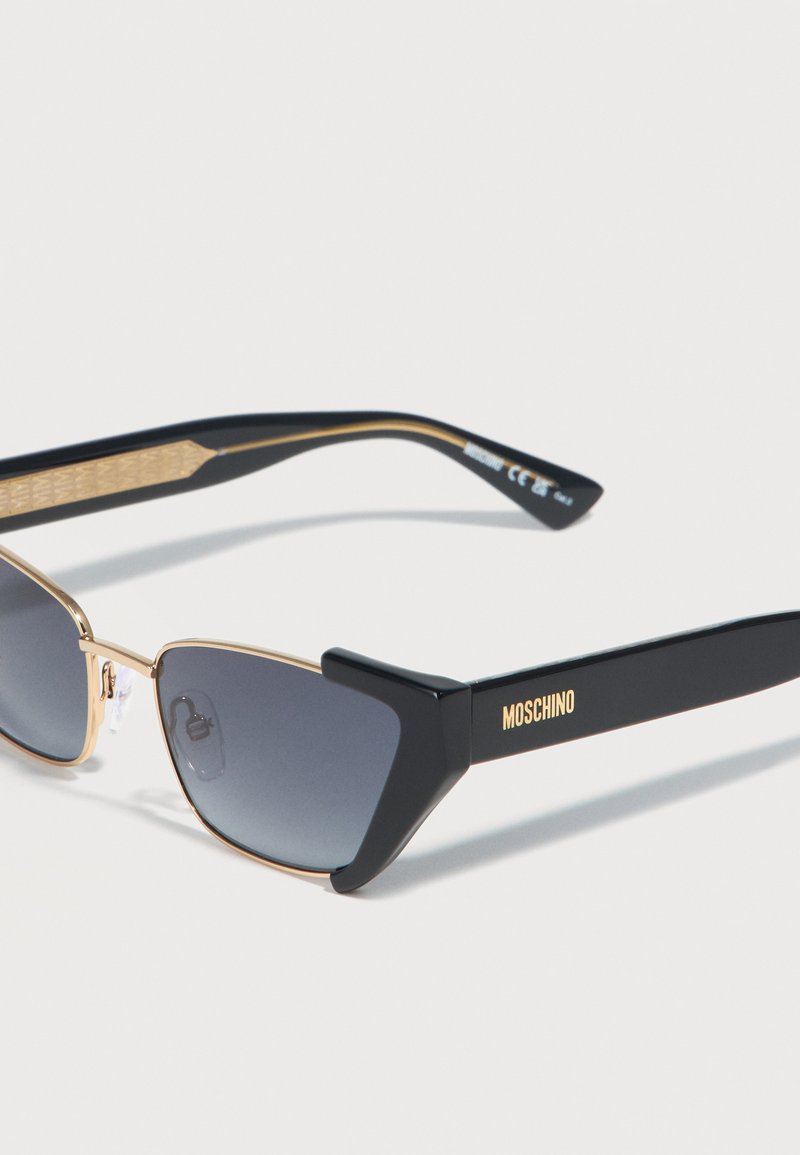 Gafas de sol Moschino en negro y dorado con lentes rectangulares oscuras y patillas negras gruesas que presentan el logotipo dorado en el lateral.