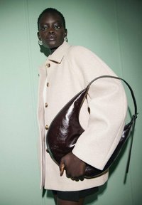 Donna di colore che indossa un cappotto beige con grandi bottoni, mentre tiene una borsa a mezzaluna lucida di colore marrone scuro contro uno sfondo verde pallido.
