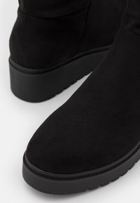 Czarne botki z sztucznej zamszowej skóry do kostki, z okrągłym noskiem, płaską gumową podeszwą i minimalnymi detalami w szyciu. Gładka tekstura i elegancki design.